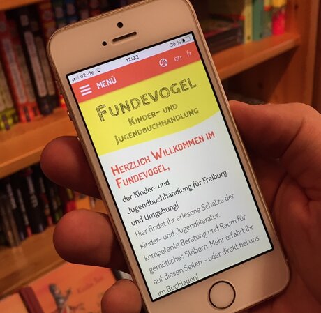 A smartphone screen shows the Fundevogel website, bookshelves in the background. - Image source: Eigenmaterial